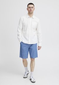 Camisa blanca de botones con un bolsillo, combinada con pantalones cortos de denim azules. El modelo lleva calcetines blancos y zapatillas negras y grises con tres rayas.