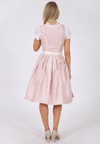 Femme portant une robe rose pâle à motifs, longueur genoux, corsage ajusté, et un chemisier blanc à manches courtes, debout de dos sur un fond uni.