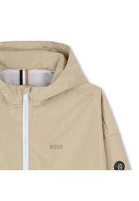 Giacca a vento beige con cappuccio, texture liscia, zip bianca e patch logo nero. Presenta dettagli a righe all'interno del collo e fodera in mesh.