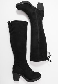 Bottes noires au-dessus du genou en suède doux. Comprend une fermeture éclair sur le côté, une surface texturée et un talon épais et haut avec une bonne adhérence.