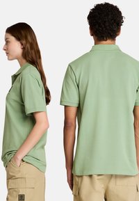 Gröna pikétröjor med korta ärmar, typisk krage och avslappnad passform visas på två modeller, en framifrån och en bakifrån.