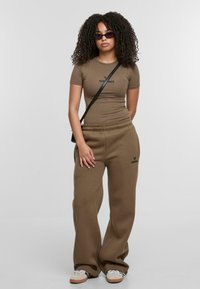 Braunes, enganliegendes Hemd mit dem Schriftzug "real", lockere braune Sweatpants und cremefarbene Sneakers. Schwarze kleine Umhängetasche und dunkle Sonnenbrille. Lockige Haare.