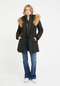 Cappotto piumino nero con cappuccio in pelliccia sintetica staccabile, chiusura con zip e tasche; indossato sopra una giacca interna nera trapuntata e jeans blu.