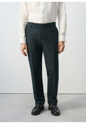 Mężczyzna ubrany w czarne spodnie w drobną prążek, czarne skórzane loafersy i białą koszulę z guzikami, stojący na jasnoszarej podłodze.