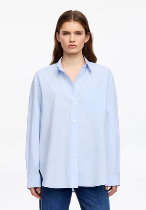 Femme aux longs cheveux bruns portant une chemise surdimensionnée bleu clair à boutons et un jean bleu, debout devant un fond blanc uni.
