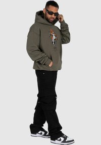 Felpa verde oliva con stampa grafica raffigurante un cavaliere, abbinata a pantaloni cargo neri e sneakers in bianco e nero.