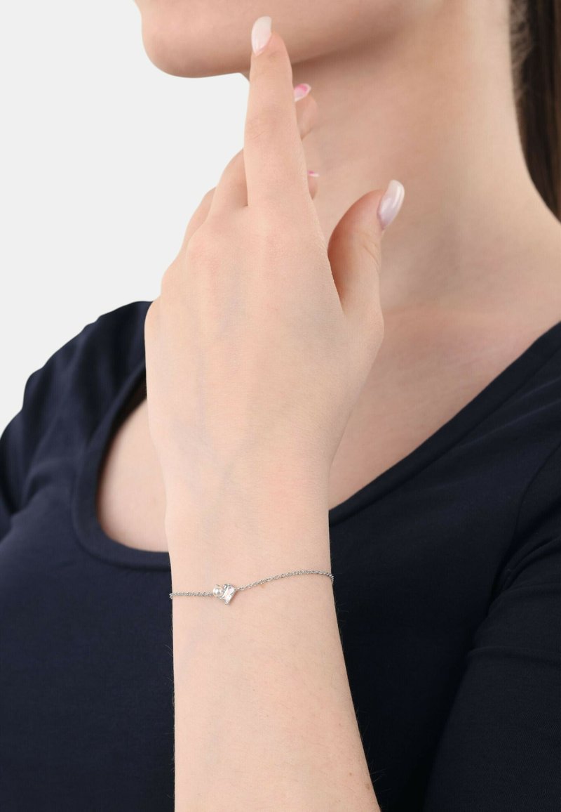 Silverarmband med en känslig kedja, prytt med en liten diamantliknande detalj, bärs på en hand mot en mörk klädbakgrund.