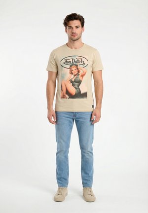 Giovane uomo in piedi che indossa una t-shirt grafica beige Von Dutch, jeans azzurri e sneakers beige, su sfondo bianco uniforme.