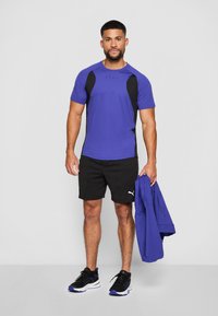 Herre treningstøy: lilla kortermet skjorte med svarte detaljer, svarte shorts, svarte joggesko, holder en blå jakke. Lettvektsstoff, sporty design.