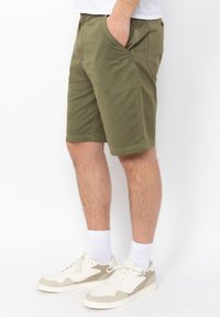 Pantalones cortos de algodón verde oliva con bolsillos laterales, un corte relajado y un detalle de botón en el bolsillo trasero. Combinados con zapatillas blancas y calcetines.
