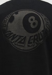 Μαύρο ύφασμα με μια σκούρα γκρι γραφική παράσταση μπάλας 8 και το κείμενο «Santa Cruz» να κυρτώνεται γύρω της με έντονα, στυλιζαρισμένα γράμματα.