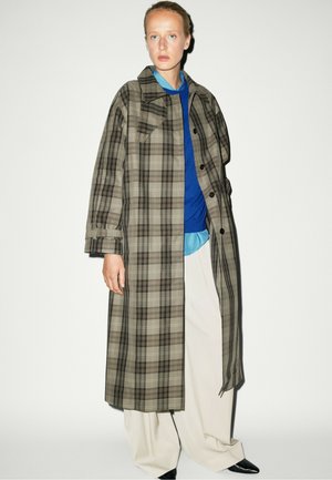 Persona che indossa un lungo cappotto a quadri marrone e grigio sopra un maglione blu, camicia azzurro chiaro, pantaloni bianchi a gamba larga e scarpe nere a punta.