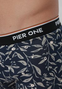 Primer plano de la cintura de una persona que lleva ropa interior azul marino con un patrón floral y una banda negra en la cintura con la etiqueta "PIER ONE" en letras blancas.