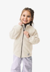 Fleecejacke in Creme mit strukturiertem Oberflächen, durchgehendem Reißverschluss und gerippten Bündchen. Tragen über einem dunklen Oberteil und hellen Hosen.
