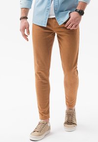 Pantalones de corte slim color tostado con una textura suave, combinados con zapatillas beis y una camisa de botones azul claro. Diseño minimalista, sin patrones visibles.