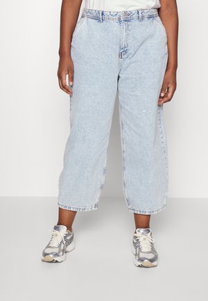 Person iført lyseblå brede cropped jeans og hvidgrå træningssko, stående mod en ensfarvet baggrund.