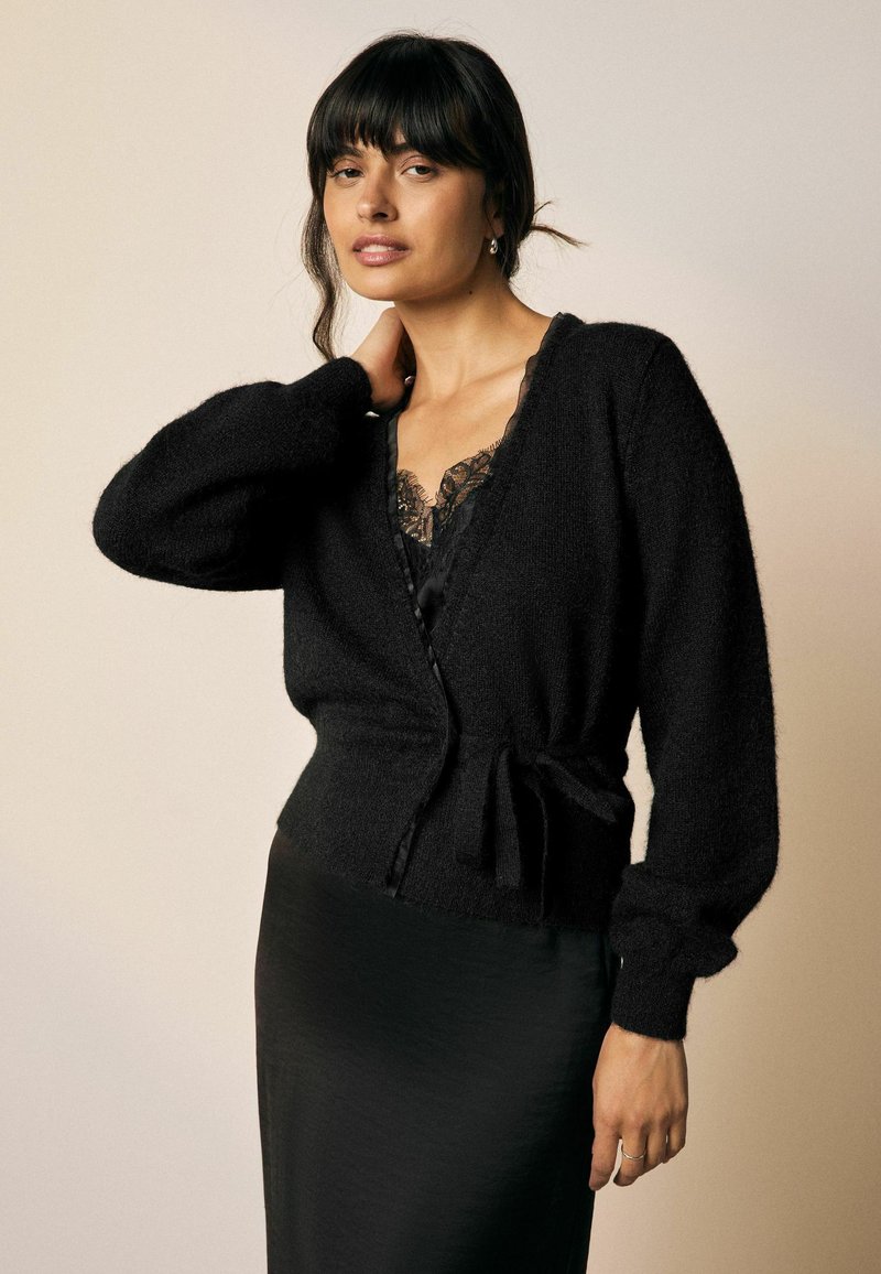 Gilet en tricot noir avec une taille ajustée, texture douce, décolleté en V plongeant et manches longues et volumineuses. Détail noué à la taille pour mettre en valeur la silhouette.