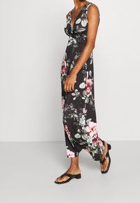 Robe longue fleurie avec un décolleté en V profond, présentant un fond noir, des motifs floraux roses et blancs, fabriquée en tissu léger. Sandales plates.