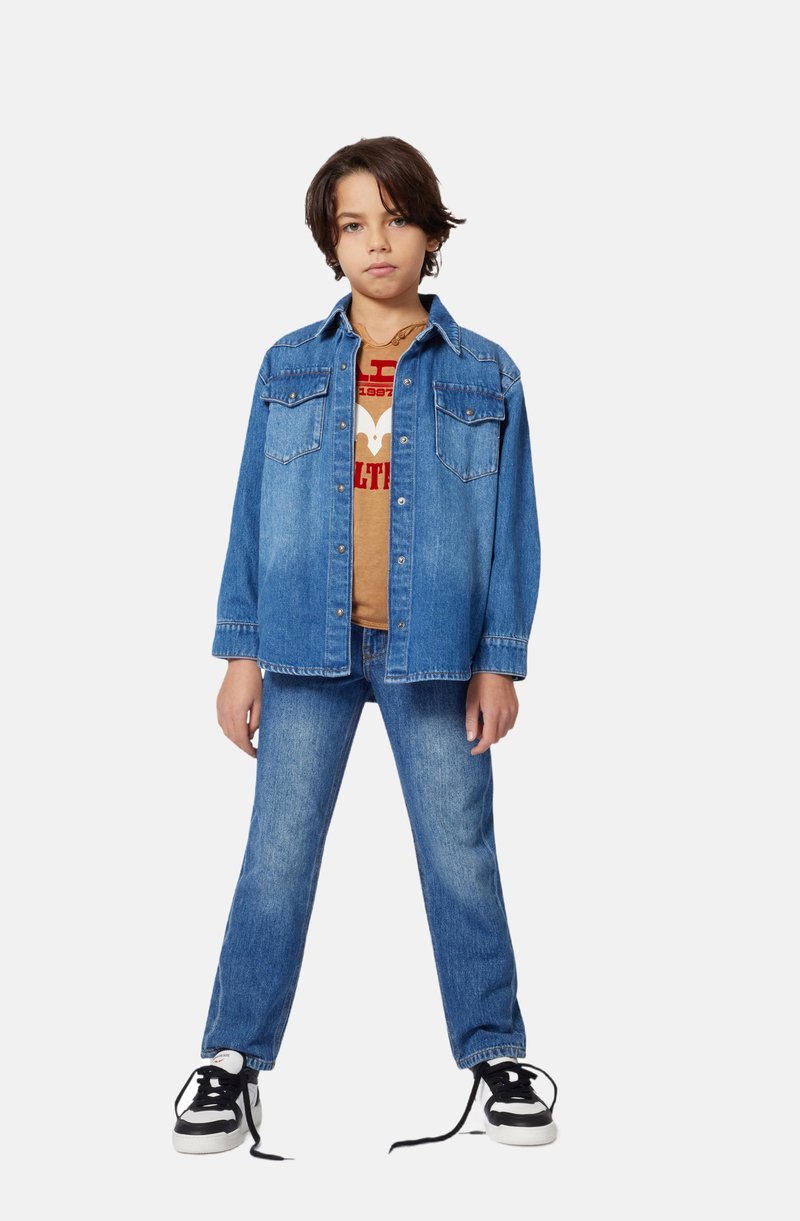 Jeansjacke mit zwei Brusttaschen, blaue Jeans und ein graues T-Shirt in Beige mit Grafik. Das Outfit wird durch schwarz-weiße Sneaker ergänzt.
