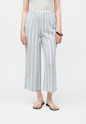 Persoon die witte broek draagt met verticale lichtblauwe strepen, beige open-toesandalen en een bruine armband aan de rechter pols.