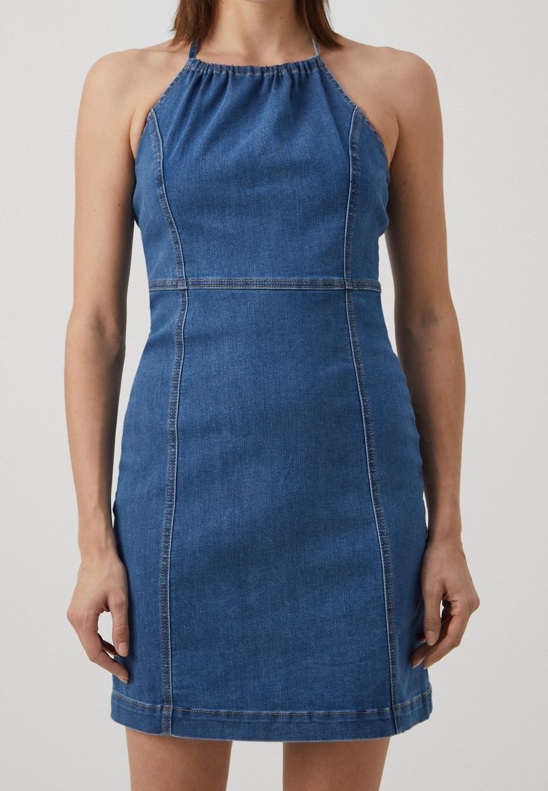 Femme portant une robe sans manches en denim bleu à col licou, debout contre un fond clair uni, les bras détendus le long du corps.