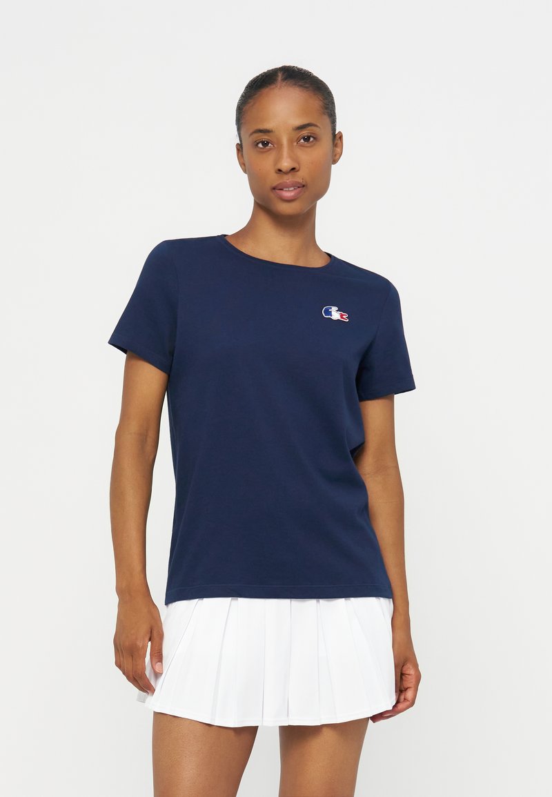 Femme portant un t-shirt bleu marine à manches courtes avec un petit logo sur la poitrine et une jupe plissée blanche, debout devant un fond uni.