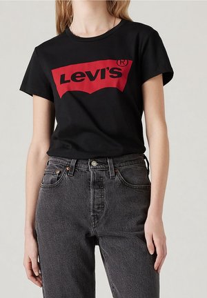 Kobieta w czarnej koszulce Levi's z czerwonym logo oraz wysokim stanem, wyblakłych czarnych dżinsach, stojąca na jednolitym tle.