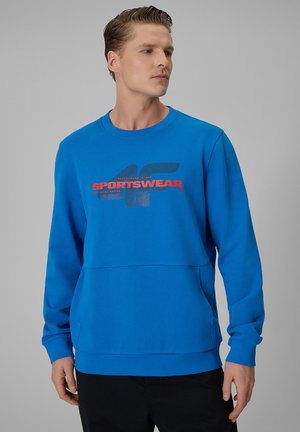 Mężczyzna noszący jasnoniebieską bluzę z logo "4F SPORTSWEAR" w czerwonym i czarnym tekście, stoi na jednolitym szarym tle.