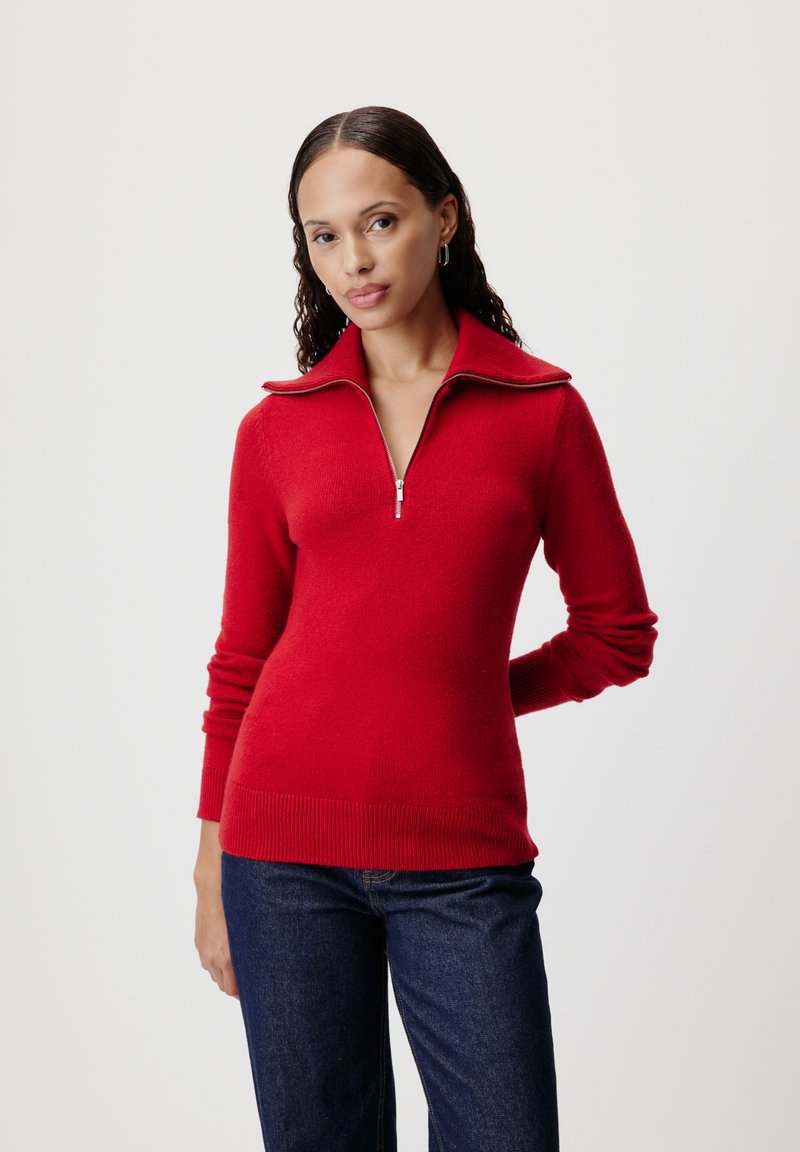 Maglione rosso con zip e colletto alto, realizzato in materiale morbido. Maniche e orlo a costine, silhouette aderente, abbinato a jeans scuri.