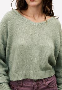 Hellgrüner, kürzer geschnittener Pullover mit V-Ausschnitt, aus strukturiertem Strickstoff gefertigt. Verfügt über überschnittene Schultern und einen gerippten Saum. Kombiniert mit lila Hosen.