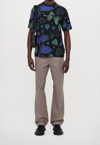 Osoba w czarnej koszuli z krótkim rękawem we wzór kwiatowy, jasnobrązowych spodniach, czarnych trampkach oraz czarnej torbie na ramię z markowym paskiem.