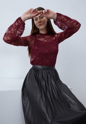 Burgundy lace long-sleeve top with floral patterns, paired with a black pleated leather skirt. Model poses with hands raised.