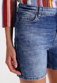 Lähikuva henkilöstä, joka on pukeutunut sinisiin kulutettuihin farkkushortseihin ja värikkäisiin raidallisiin paitaan, jossa on kolmen neljänneksen hihat.