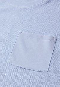 Primo piano di un tessuto azzurro chiaro che mostra una tasca quadrata cucita sul petto di un indumento lavorato a maglia con trama texturizzata.