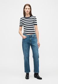 Γυναίκα που φοράει μαύρο-άσπρο ριγέ κοντομάνικο τοπ, μπλε τζιν παντελόνι με ίσιο ύφασμα και μαύρα μποτάκια αστραγάλου, στέκεται με το ένα χέρι στην τσέπη.