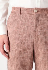 Pantalons tissés texturés dans une teinte brun rougeâtre avec une fermeture à bouton et une coupe ajustée. Le tissu présente un motif léger moucheté.