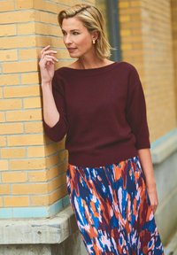 Pull en maille bourgogne avec un décolleté large associé à une jupe maxi fluide et à motifs ornée de designs abstraits en orange, bleu et blanc.