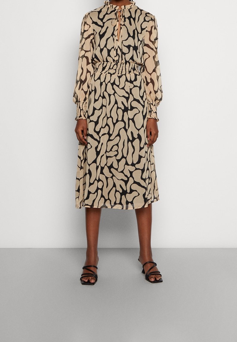 Femme portant une robe longue jusqu'aux genoux à manches longues avec un motif abstrait beige et noir et des talons noirs ouverts à brides.