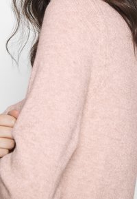 Suéter de punto rosa claro, con cuello redondo, de textura suave y con mangas largas. El material parece cálido y acogedor, con un ligero detalle de canalé.