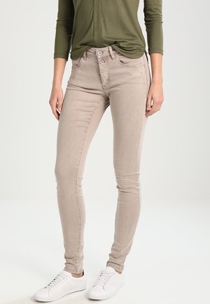 Person iført beige skinny bukser med dobbelt knaplukning, olivengrøn langærmet top og hvide sneakers, stående på en ensfarvet lys baggrund.