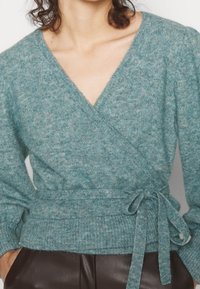 Turkos omslagströja i mjukt, texturerat stickat tyg. Har en djup V-hals, långa ärmar och ett knytbälte i midjan.