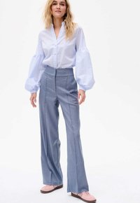 Lichtblauwe gestreepte blouse met volumineuze mouwen en knoopdetails, gecombineerd met hoog aangesloten grijze broek en lichtroze schoenen.