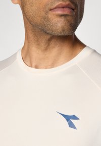 Close-up of a man's lower face and neck wearing a cream sports shirt with a small blue logo on the chest.