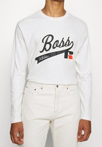 Vit långärmad skjorta med "Boss" tryckt i svart och röda accenter. Gjord av mjukt tyg, har rund halsringning och avslappnad passform.