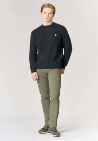 Maglione a trecce grigio scuro con scollo a girocollo, abbinato a pantaloni slim fit verde oliva e sneakers verde/grigie. Logo ricamato semplice sul maglione.