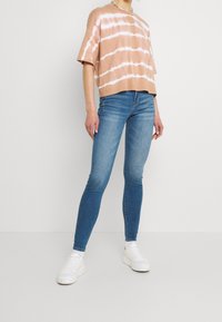 Kortärmad, avslappnad ljusbrun tie-dye bomullstopp som är ihopparad med tighta blå jeans och vita sneakers på en neutral bakgrund.