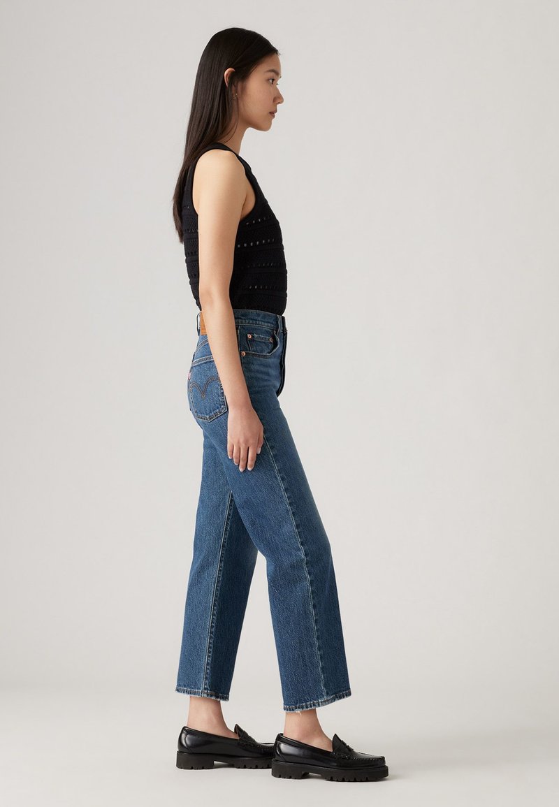 Débardeur noir en tricot, jeans bleu taille haute à coupe droite avec fentes sur les côtés, associés à des chaussures noires à enfiler. Textures lisses, coupe décontractée.