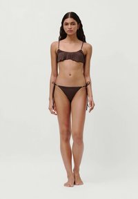 Woman standing barefoot against a plain background wearing a brown bikini with ruched top and tie-side bottoms.