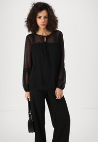 Blusa nera con maniche lunghe a sbuffo in trasparenza, con pois a rilievo e scollatura con nodo. Abbinata a pantaloni larghi neri e una borsa piccola.