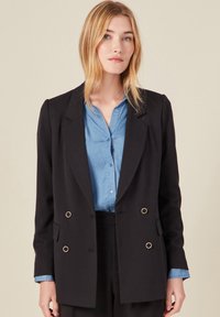 Blazer noir à double boutonnage avec des boutons en métal doré, revers crantés et une coupe ajustée, superposé à une chemise en chambray bleu.